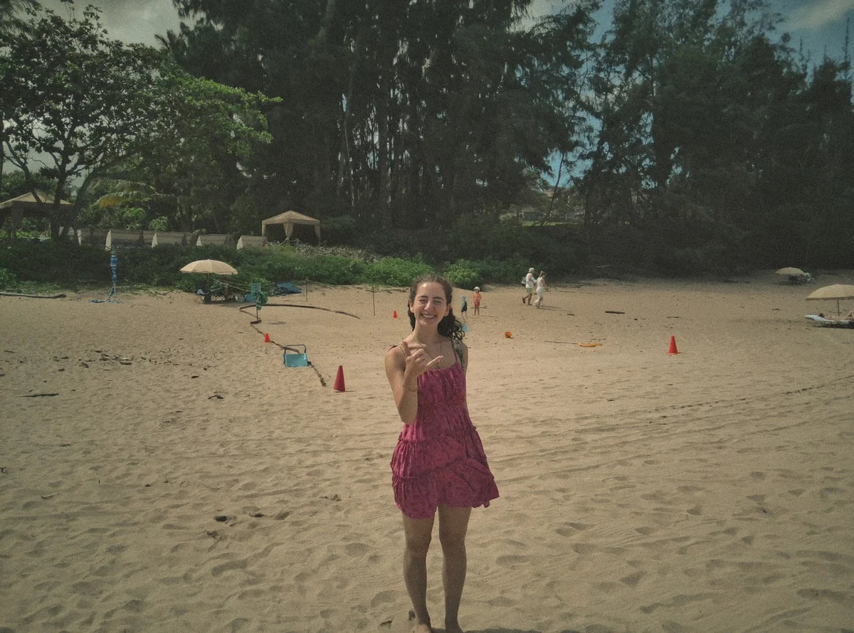 A woman smiling and making a shaka gesture on a sandy beach.