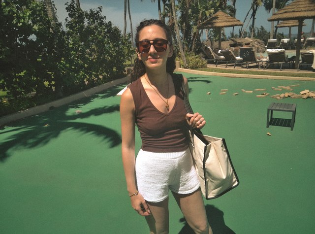 A woman wearing sunglasses and carrying a tote beside a palm-lined resort lawn.